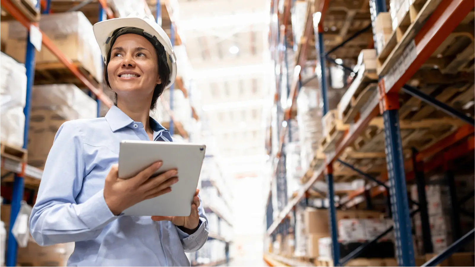Worker in a warehouse holding a tablet while standing between storage racks.